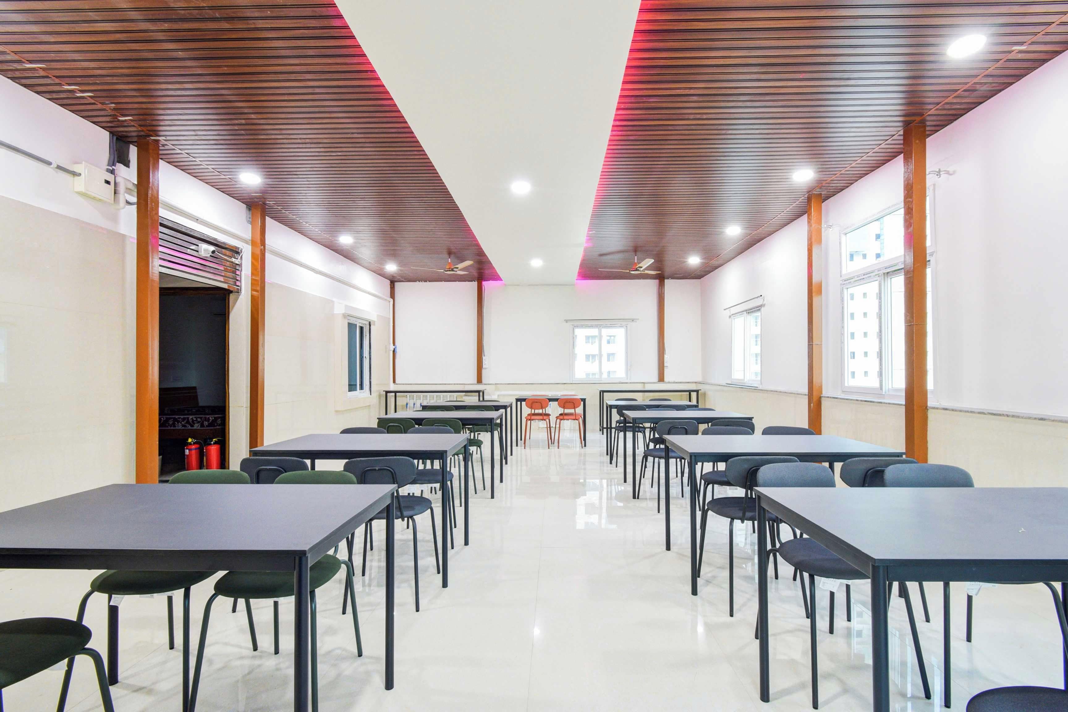 Uma sala de aula com fileiras de mesas e cadeiras, uma lousa e ventiladores de teto.