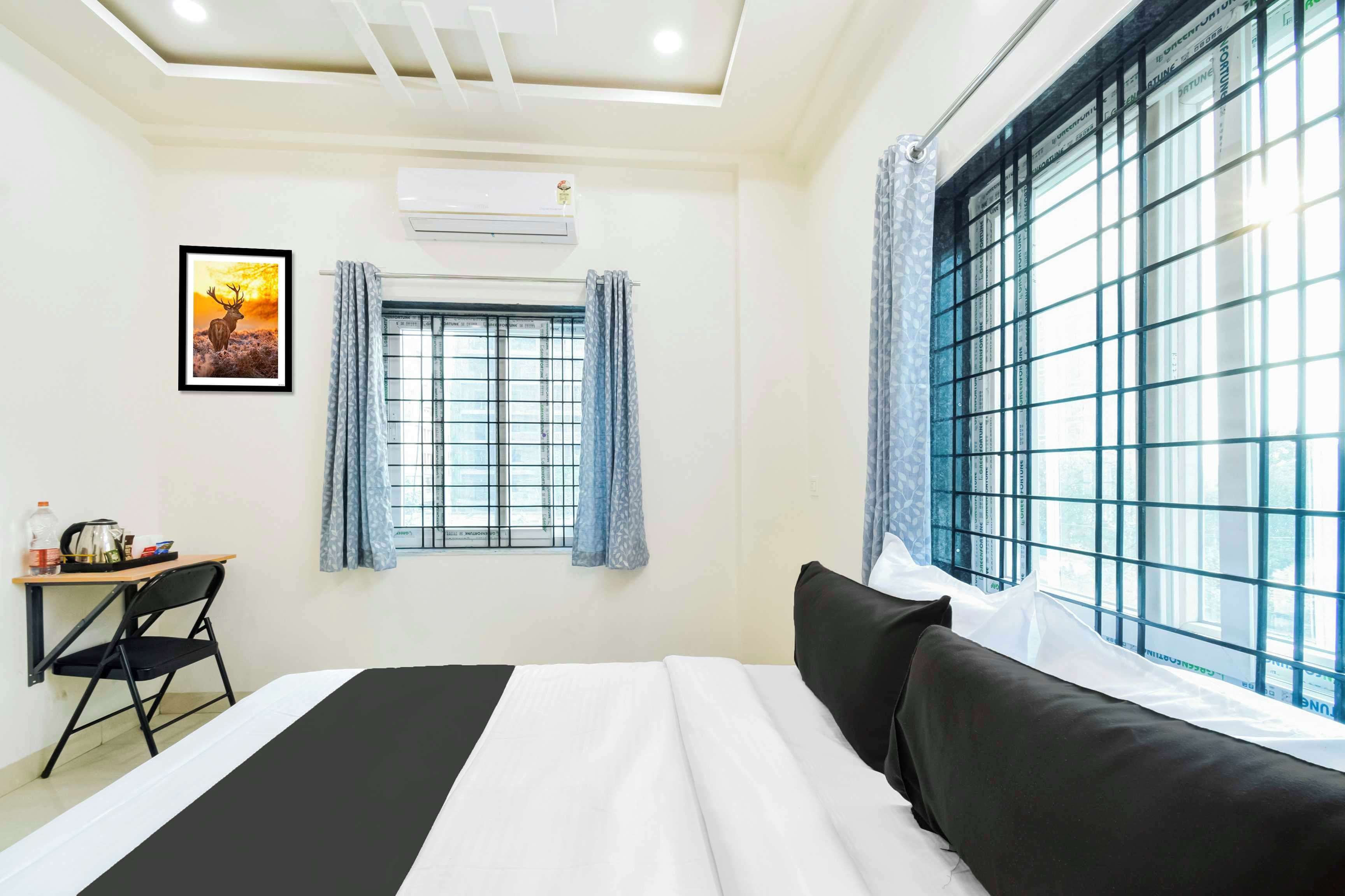 Quarto de hotel com cama, escrivaninha, cadeira, janela com cortinas e ar condicionado.