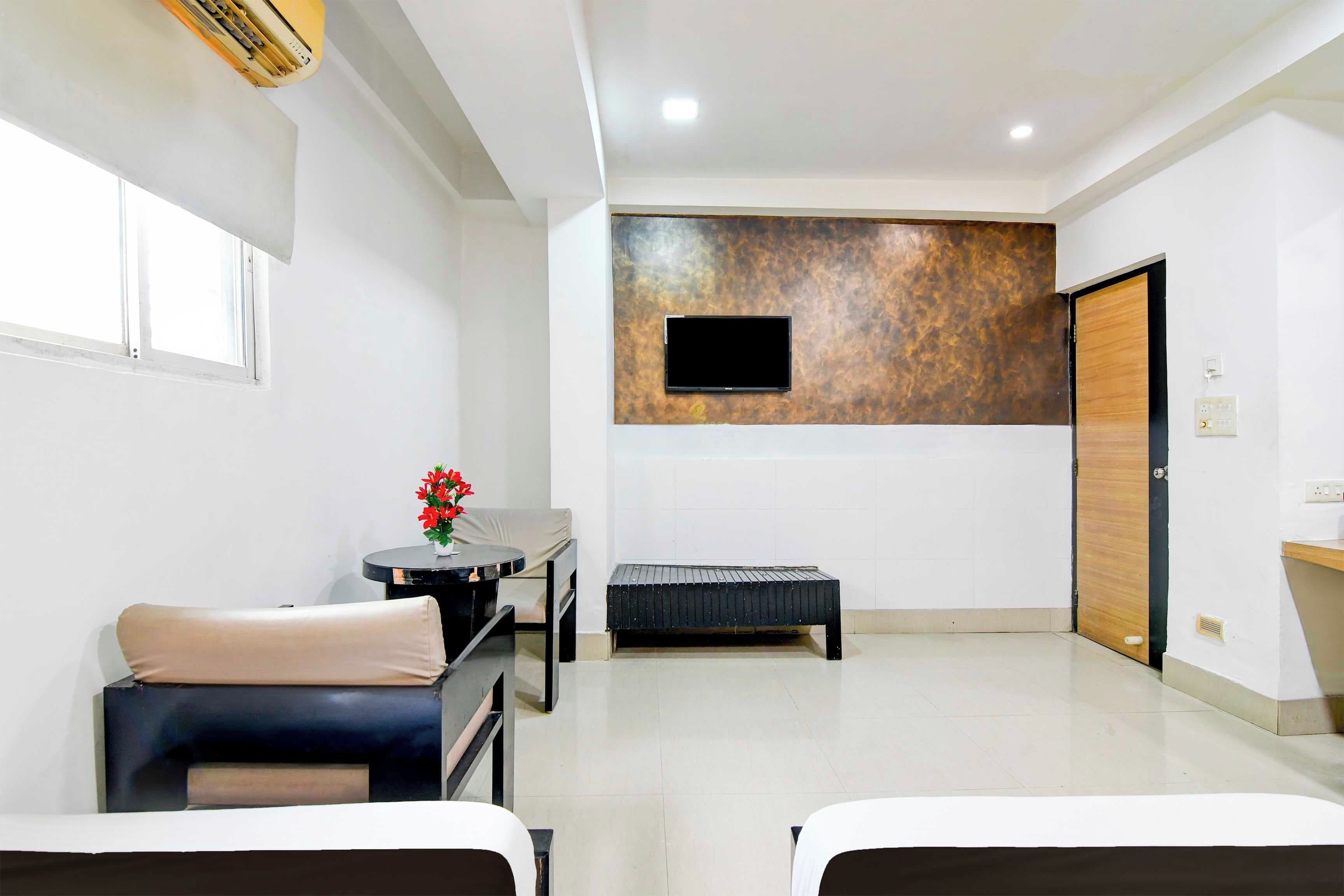 Habitación de hotel con dos camas, una mesita con un florero, una banca, un televisor instalado en una pared con textura y una puerta de madera.