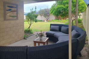 Ein überdachter Patio-Bereich mit einer dunklen Rattan-Couch, einem kleinen Holztisch mit einem Blumentopf und einer an der Wand befestigten Fischdekoration.