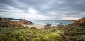 Paisagem costeira com afloramentos rochosos, uma praia arenosa e vegetação arbustiva densa.