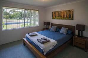 Quarto com uma cama grande, duas mesas de cabeceira com abajures, uma janela com persianas e uma pintura com tema de floresta na parede.