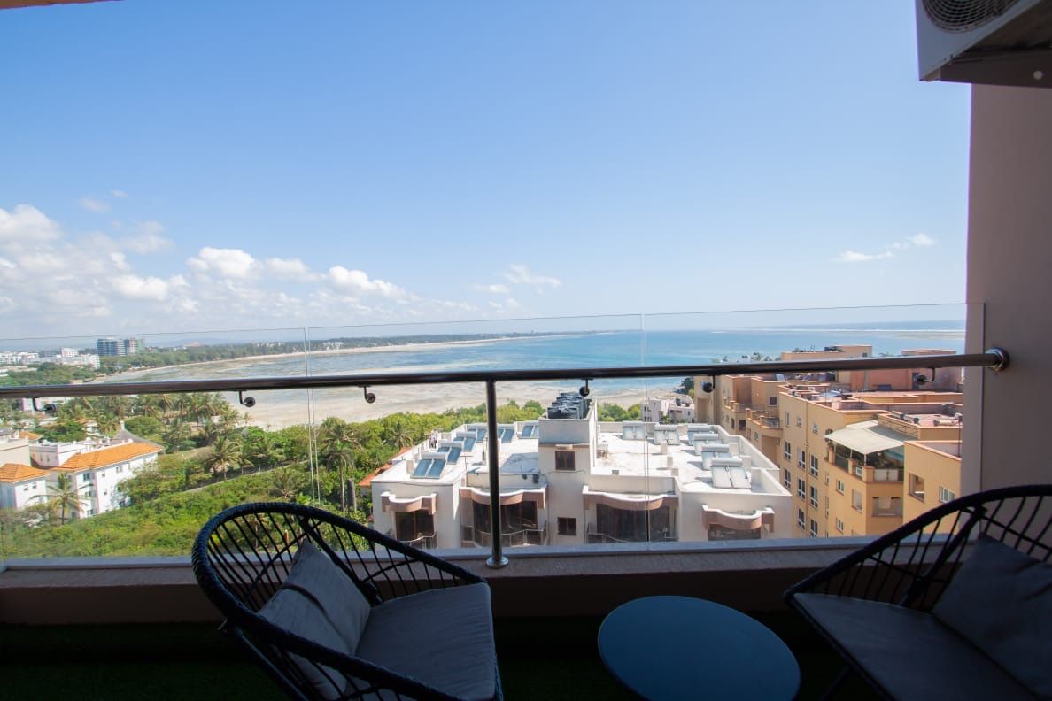 Un balcon avec deux chaises et une petite table, donnant sur une plage et des immeubles.