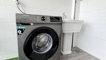 Eine kompakte, moderne Waschmaschine mit Bedienfeld, einer Steckdose und einem weißen Waschbecken in einem sauberen, gefliesten Badezimmer.