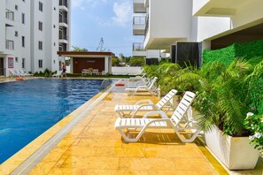 Piscina com espreguiçadeiras e placa 'Lounge', rodeada por vegetação e um edifício de vários andares.