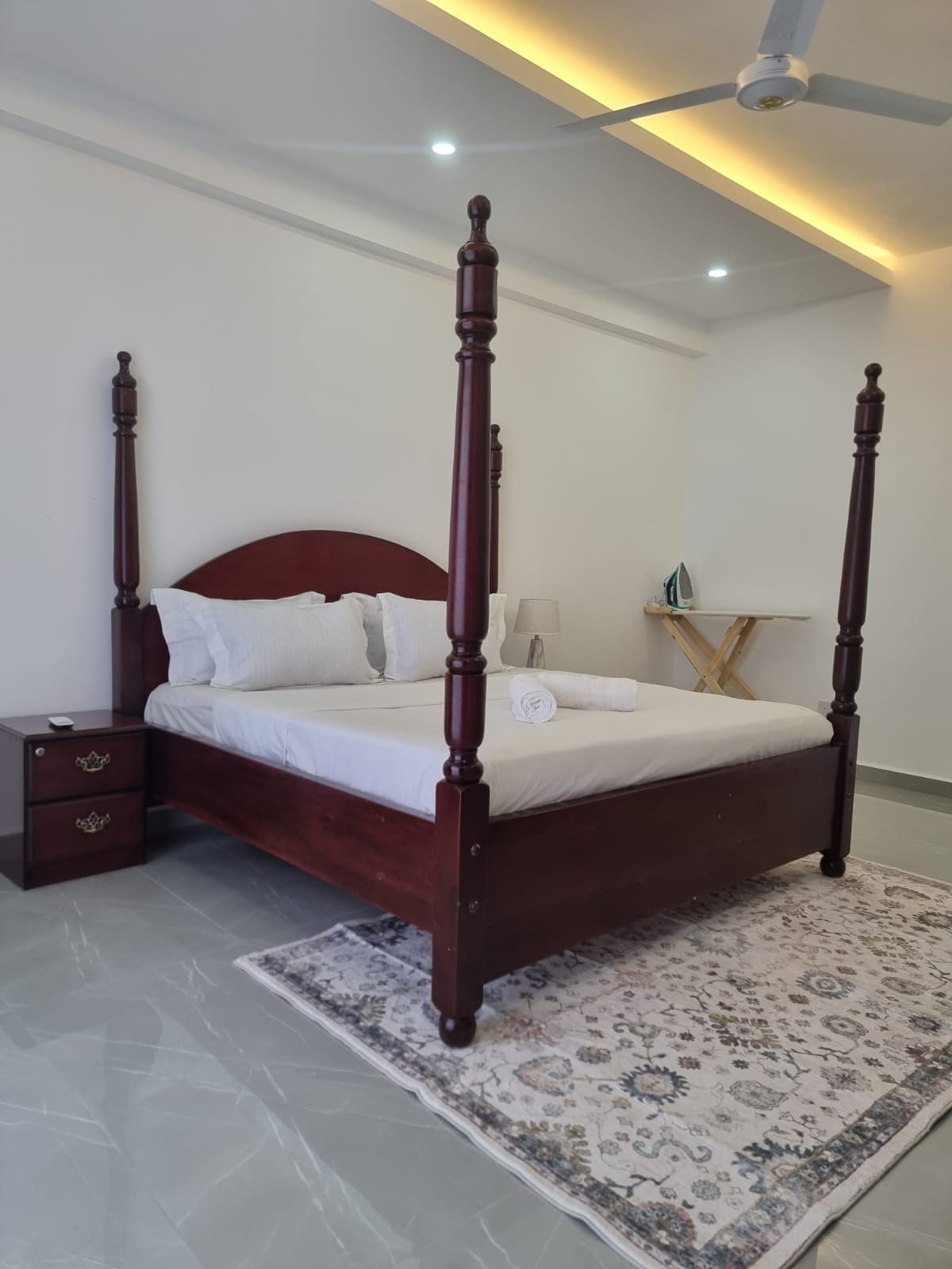 Un lit à baldaquin avec une tête de lit et un pied de lit en bois, une table de chevet avec des tiroirs, une lampe et un ventilateur de plafond.