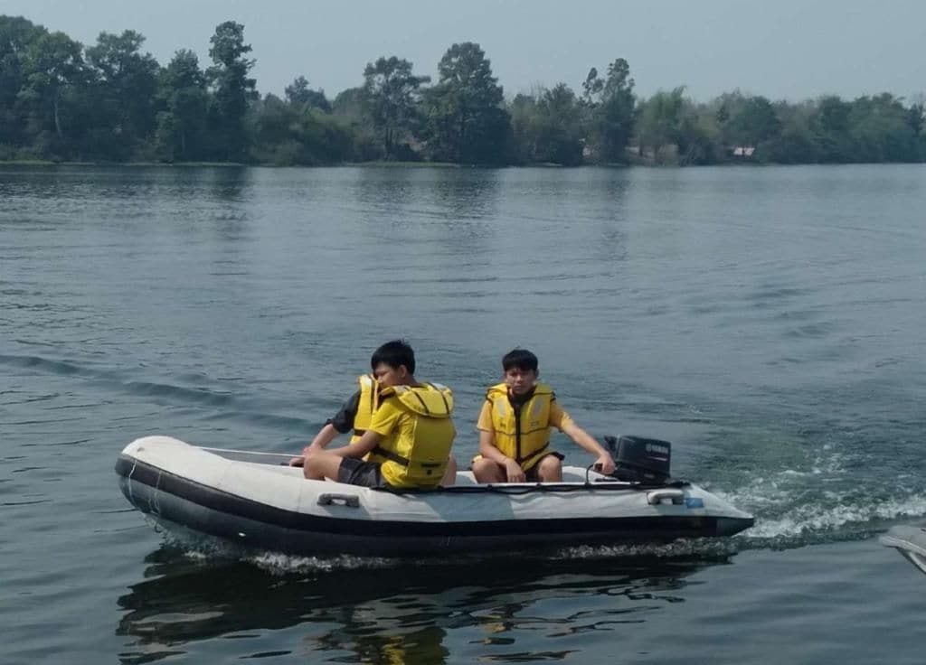 Twee mensen in een klein motorbootje, met veiligheidswesten, op kalm water met een bebost landschap op de achtergrond.