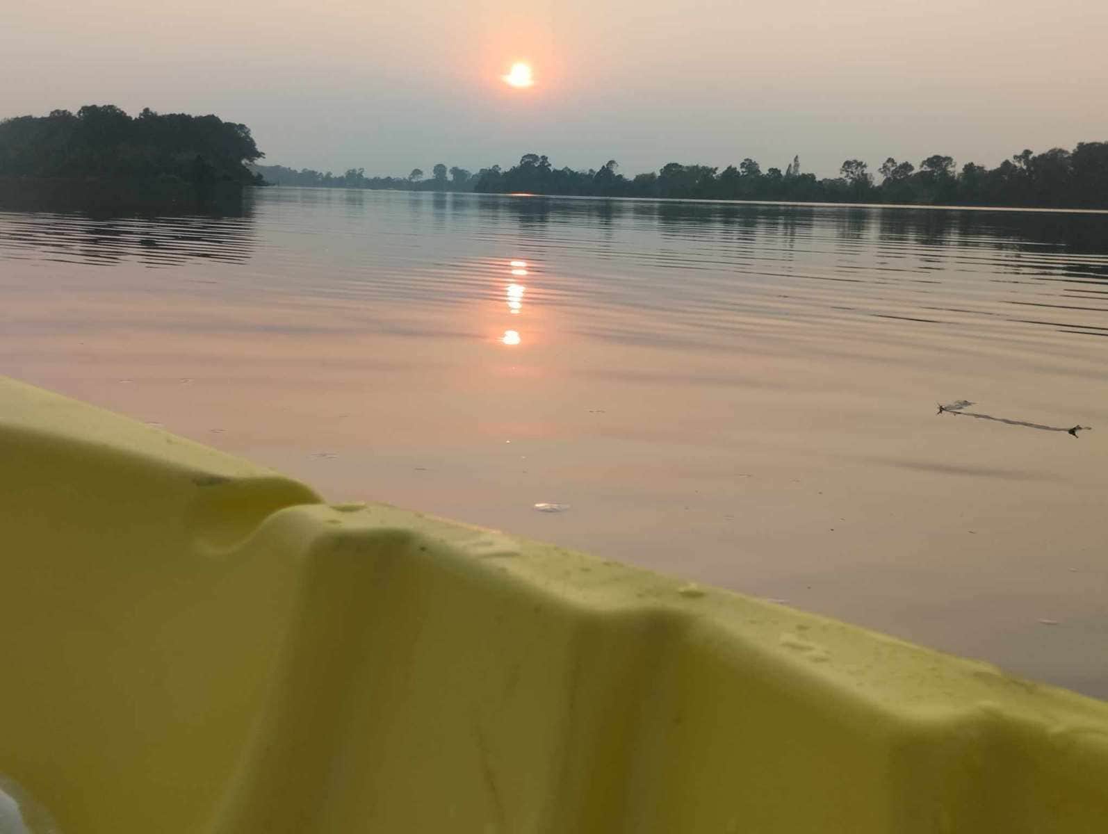 A calm body of water with a sun reflecting on the surface, surrounded by trees and a distant landmass.