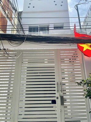 A white gate with horizontal slats, a red flag with a yellow star, and a building with a white facade and windows in the background. - Oula Home - Silk Studio l Central D1, 5 Mins to Ben Thanh Market (Thành phố Hồ Chí Minh)