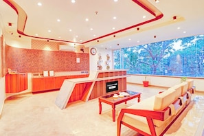 Un espace d’accueil moderne avec un comptoir de réception en bois, un coin salon avec un canapé et des fauteuils, une grande fenêtre donnant sur les arbres et une horloge au mur.