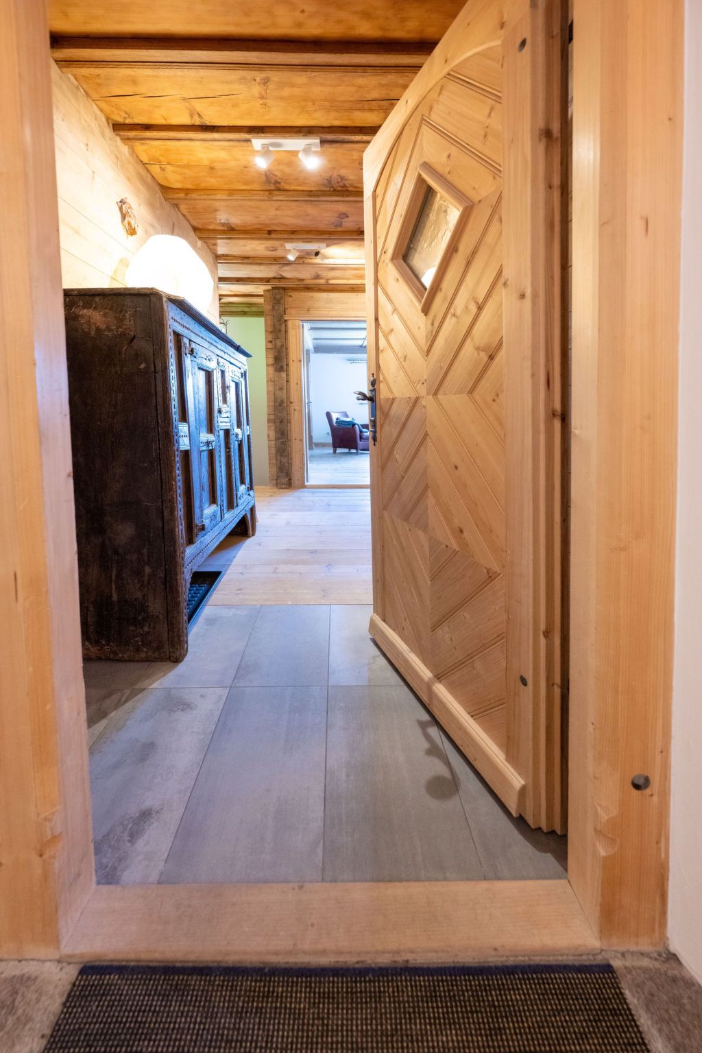 A wooden hallway with a radiator, a door with a geometric pattern, and a glimpse of a room with a chair and a table.