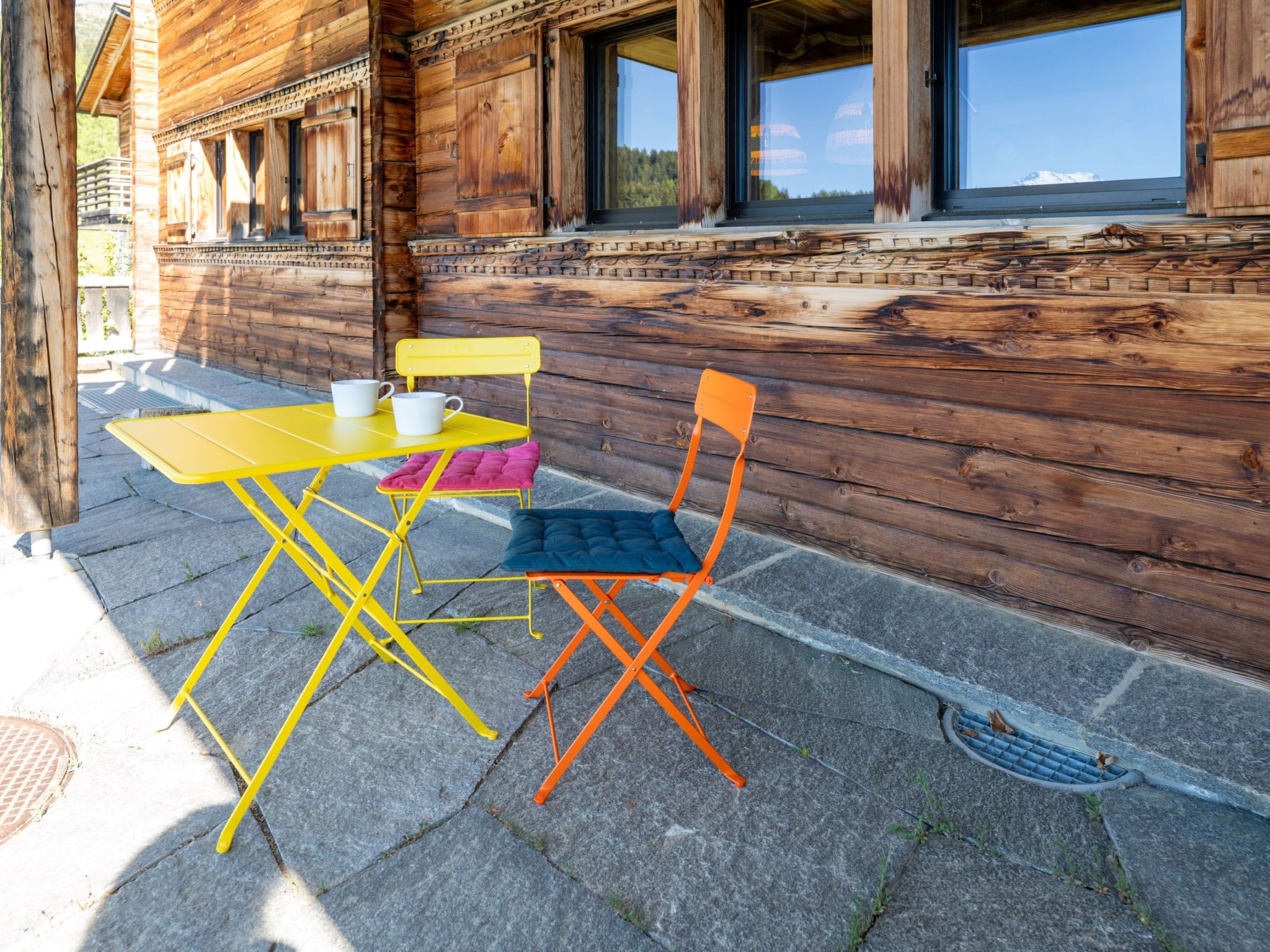 A wooden cabin with a patio table and two chairs, a window, and a view of the mountains.