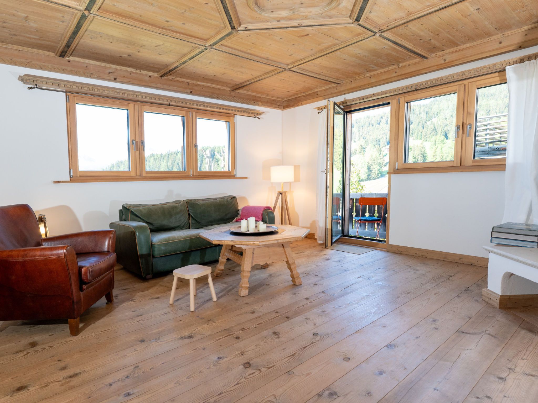 Ein gemütliches Zimmer mit Holzbalkendecke, einem grünen Sofa, einem Holztisch mit Kerzen und großen Fenstern mit Blick ins Freie.