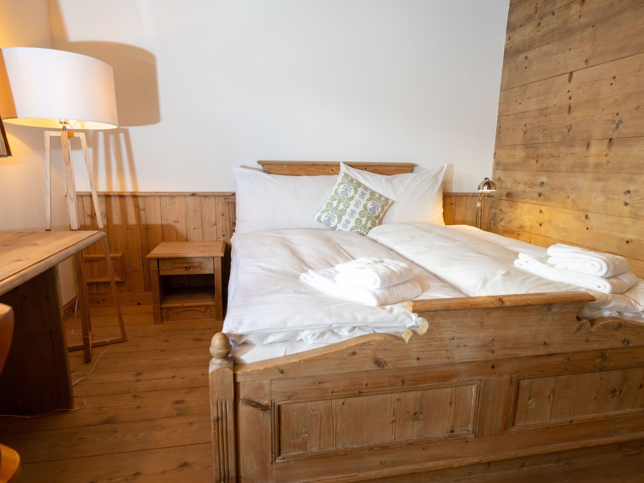 A wooden bed with white linens and towels, a bedside table, a lamp, and a wooden wall.