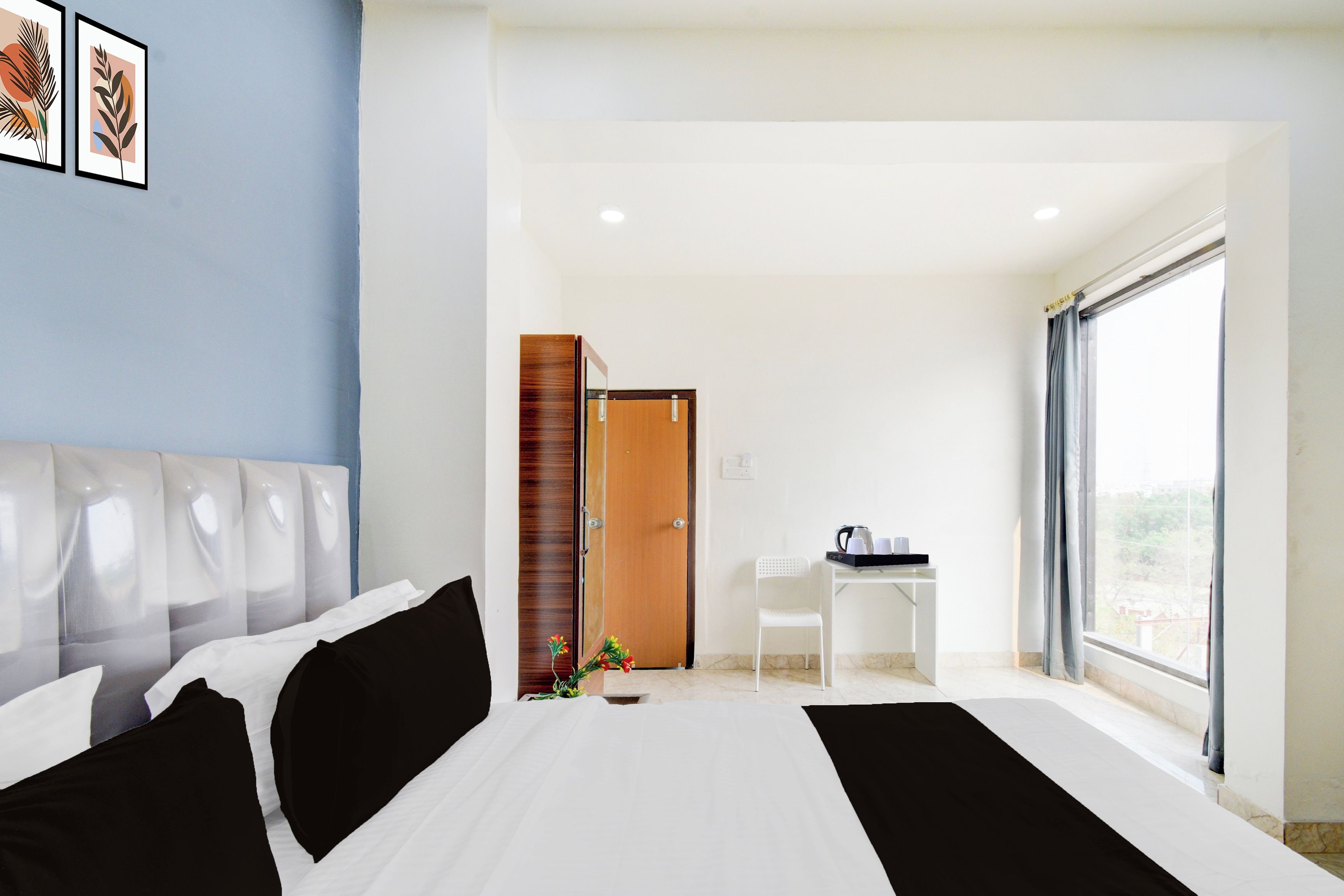 A hotel room with a bed, a desk with a chair, a window, and artwork on the wall.