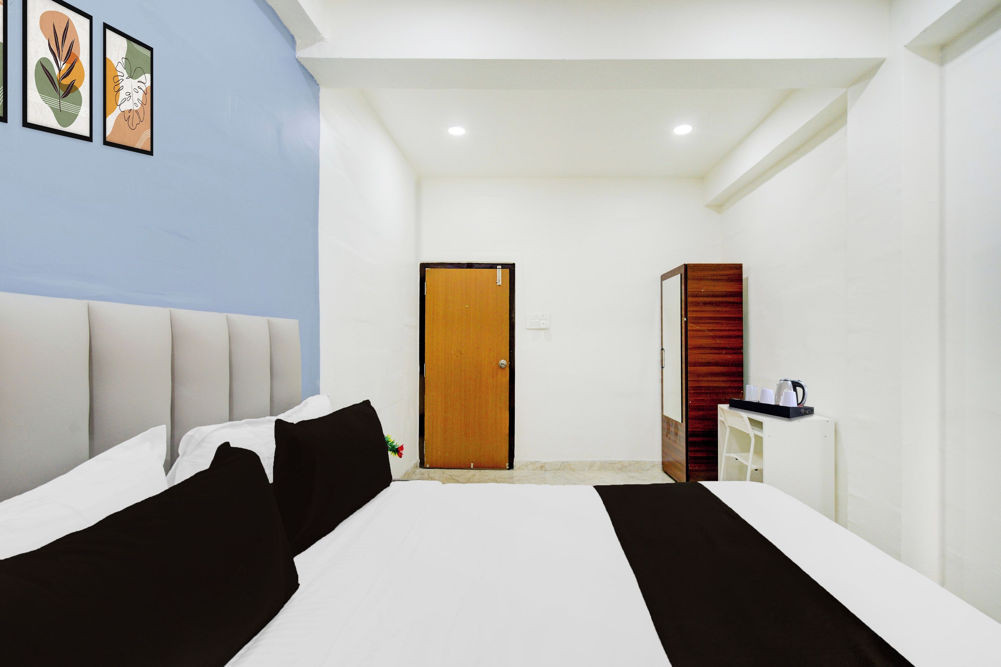 Habitación de hotel con una cama, una puerta de madera, un escritorio blanco con cafetera y cuadros en la pared.