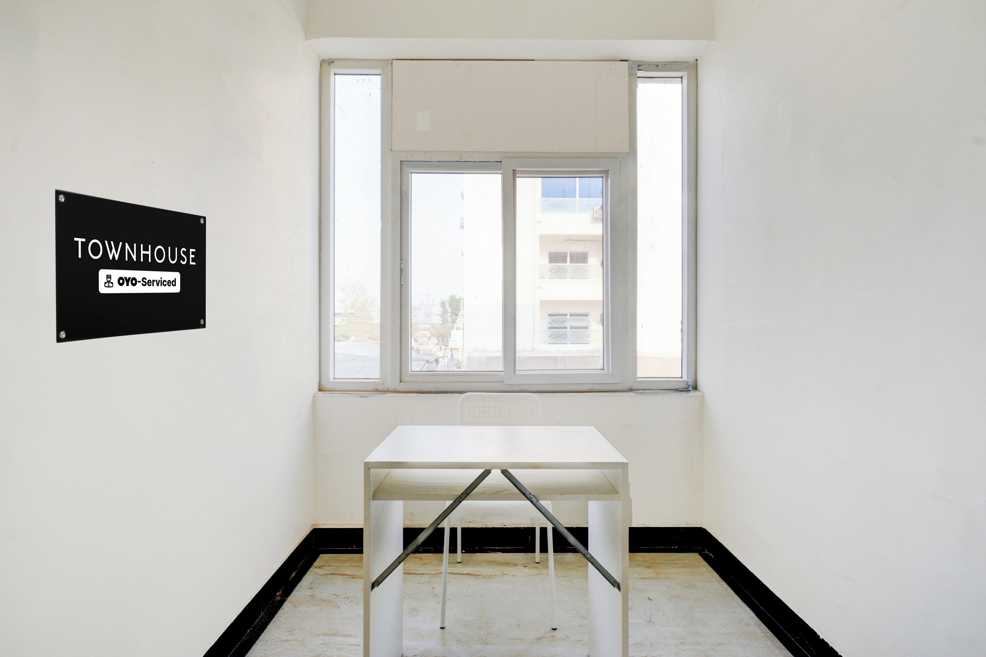 Een kamer met een witte tafel en een raam. Op de muur hangt een bordje met de tekst 'TOWNHOUSE OYO-Serviced'.