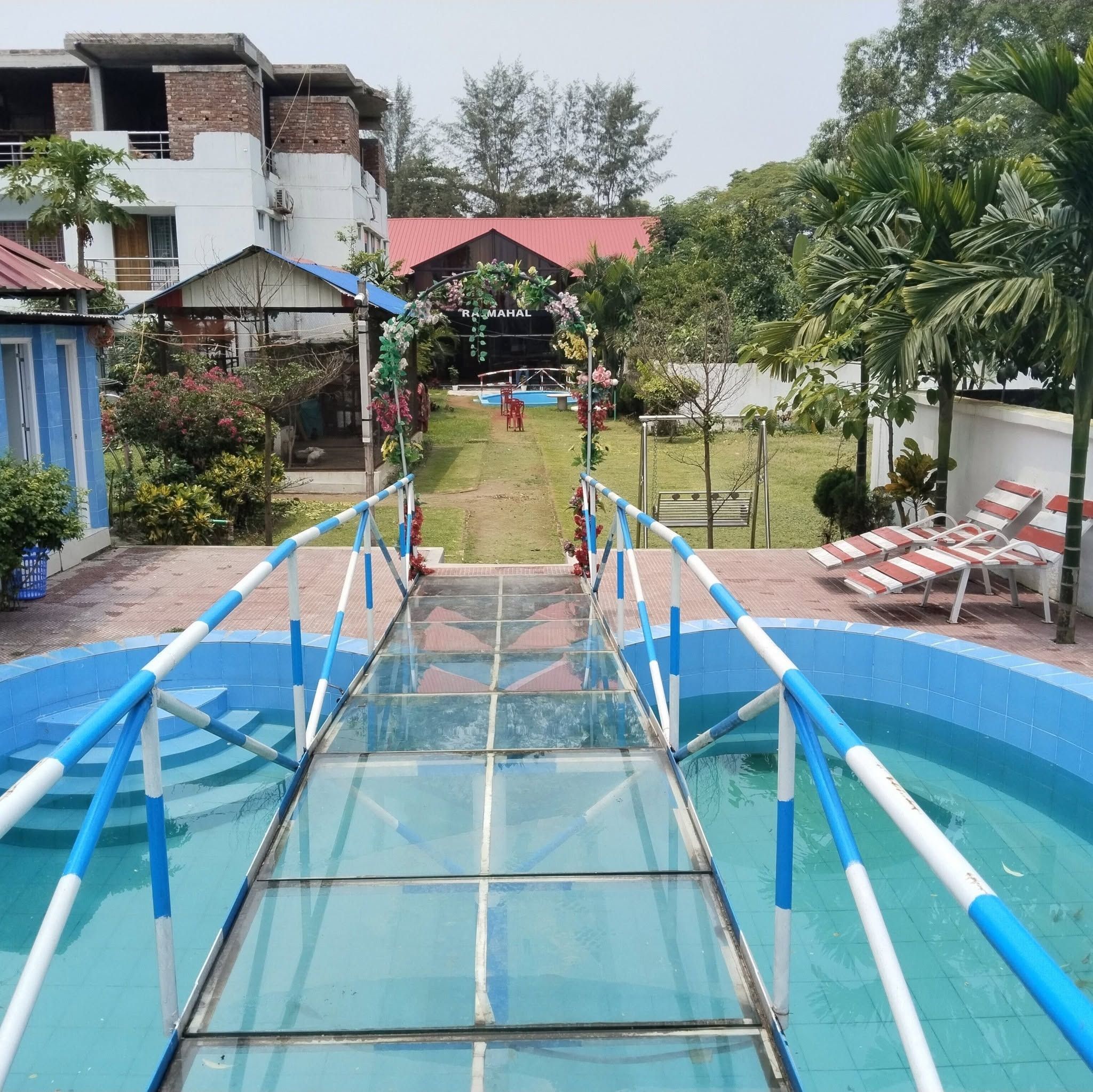 Piscina com uma ponte de vidro que leva a uma área gramada com um edifício de telhado vermelho.