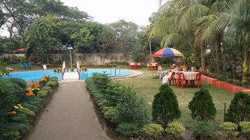 Área de piscina com caminho, assentos e uma pessoa sob um guarda-sol colorido.