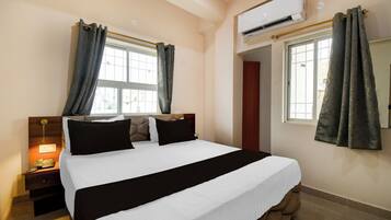 A hotel room with a bed, a nightstand, an air conditioning unit, and a window with curtains.