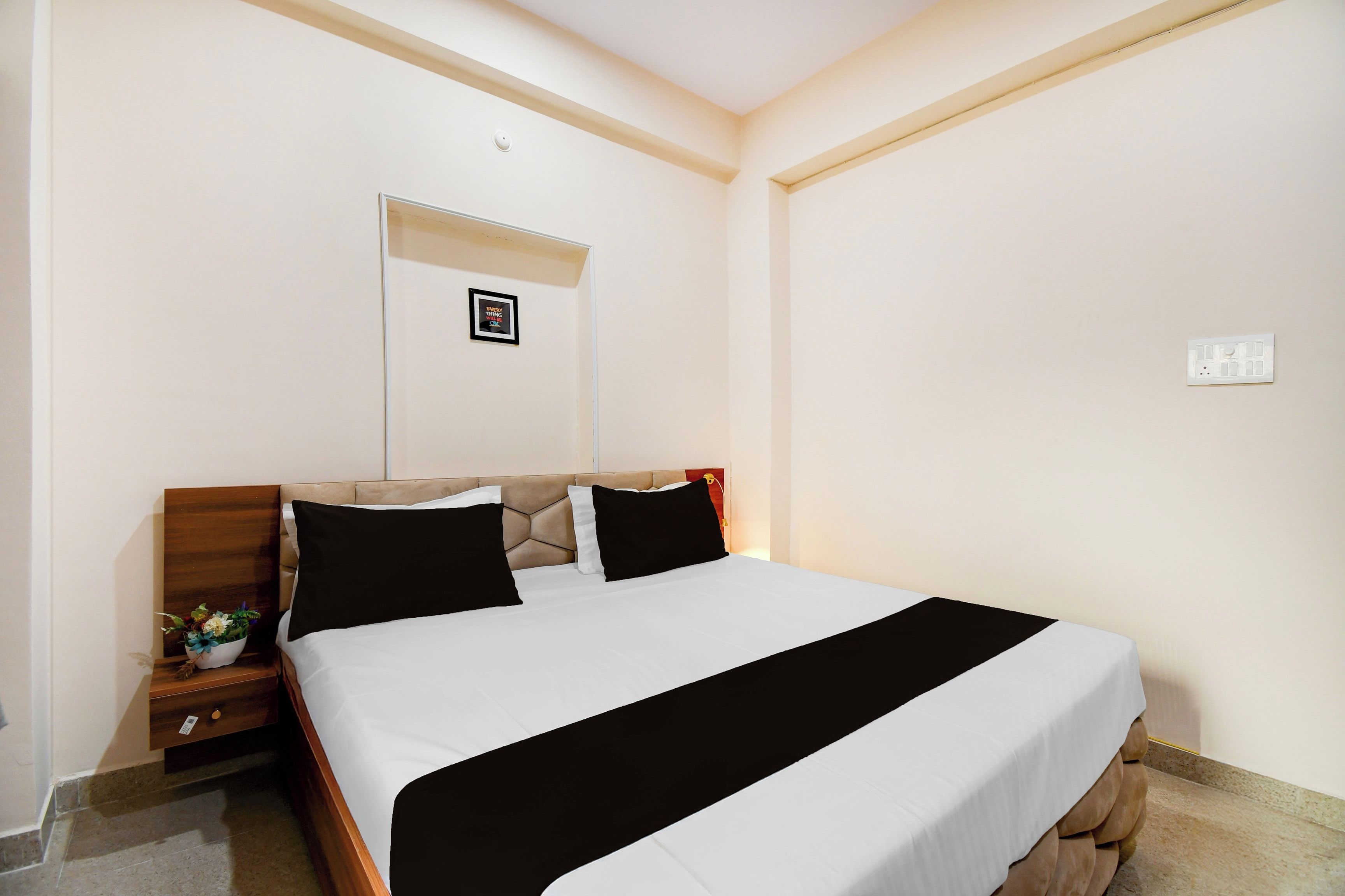 Habitación de hotel con cama, mesita de noche, una pequeña mesa con una planta y un interruptor de luz en la pared.