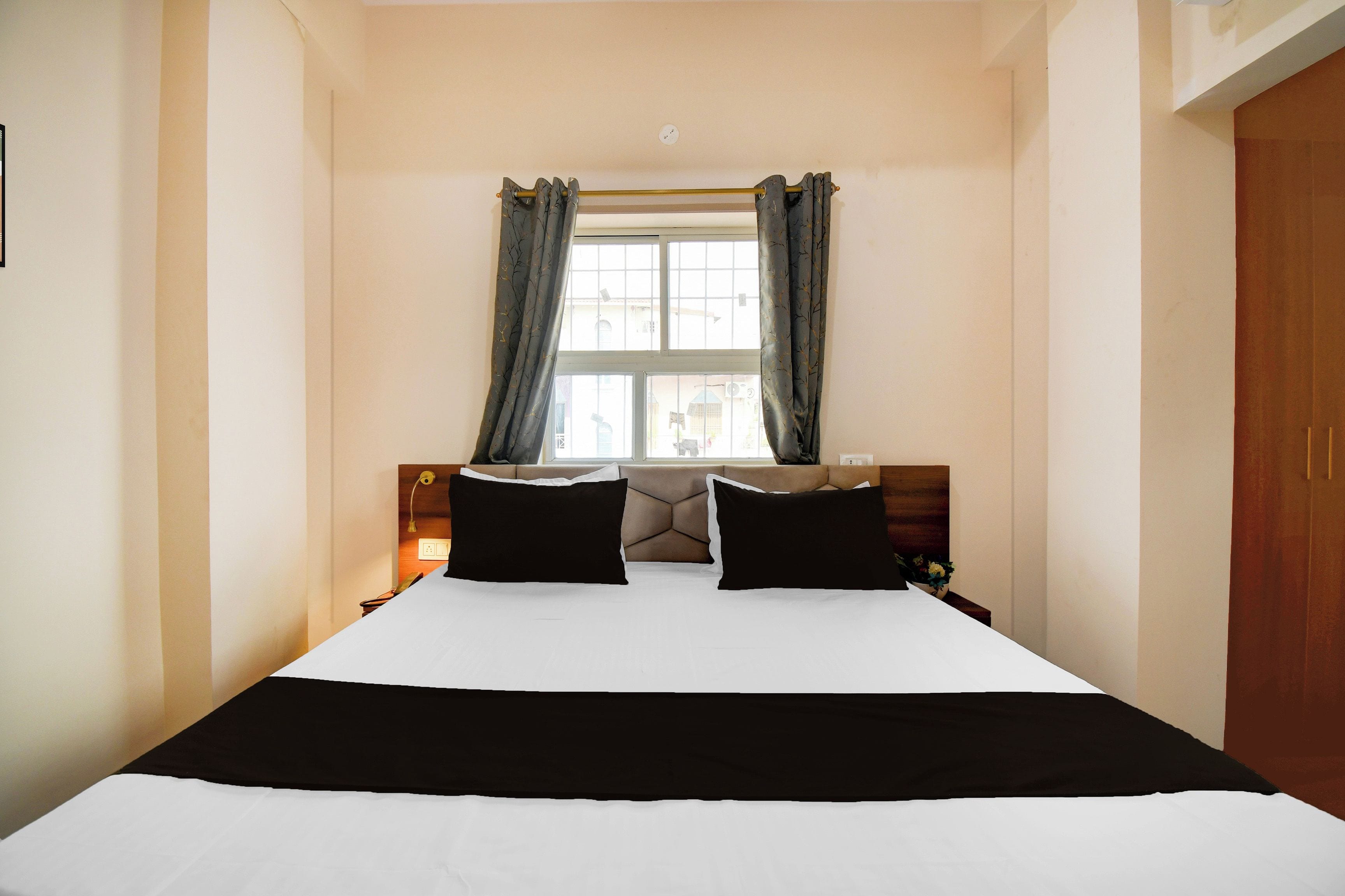 Habitación de hotel con cama, dos almohadas negras, una colcha blanca, ventana con cortinas y cabecero de madera.