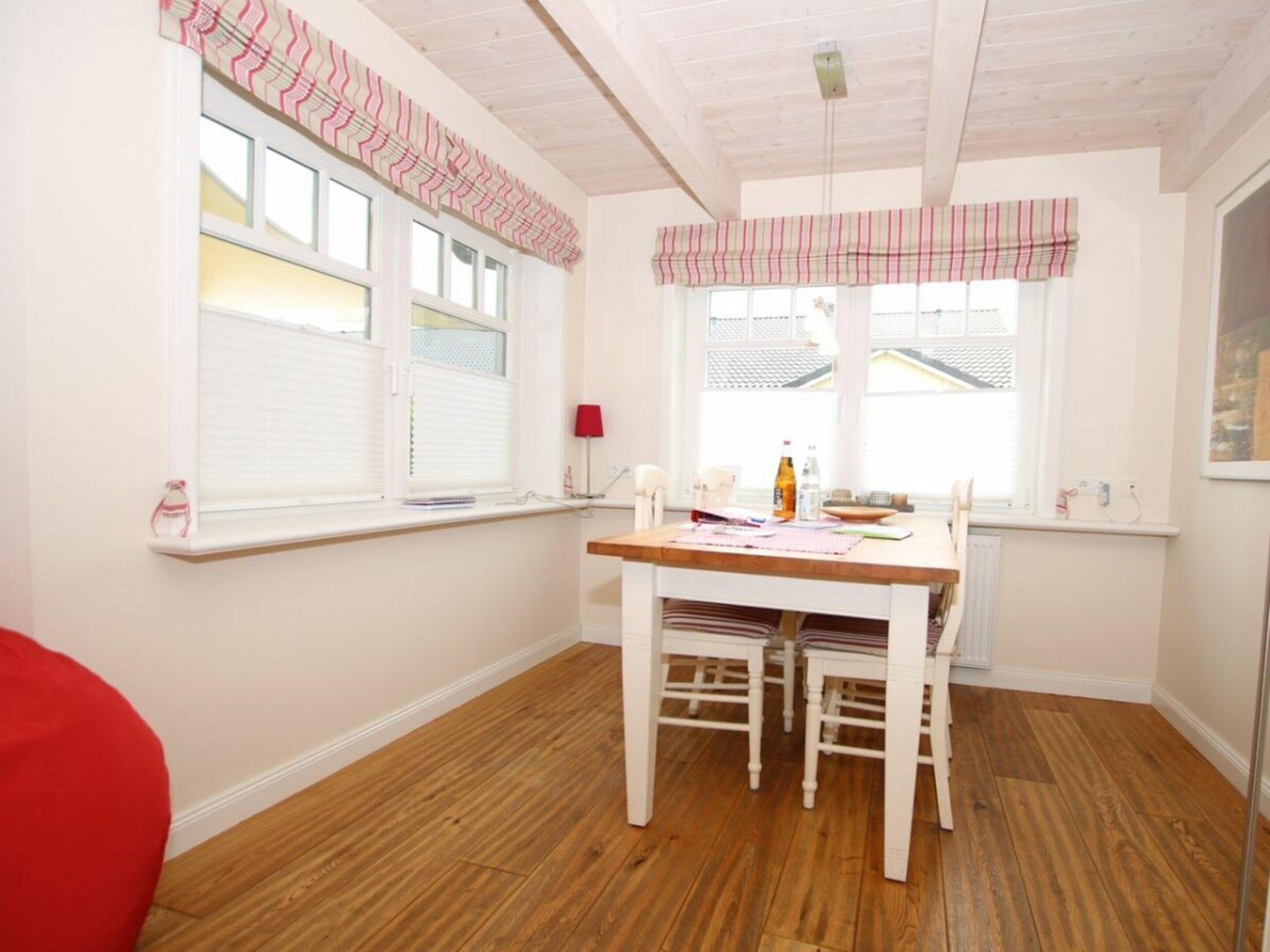 Une pièce avec un plancher en bois, une table blanche avec des chaises, une fenêtre avec des stores et une lampe rouge fixée au mur.