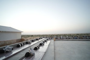 A rooftop terrace with a row of black and white striped cushions, a white tent-like structure, and a desert landscape in the background. - Jaisalmer Sand Dunes Luxury Camp and Resort (Jaisalmer)