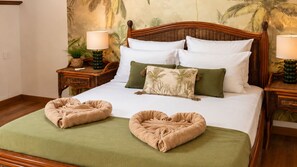 Una cama bien hecha con una colcha verde, dos toallas con forma de corazón y un cojín con estampado de hojas de palmera.