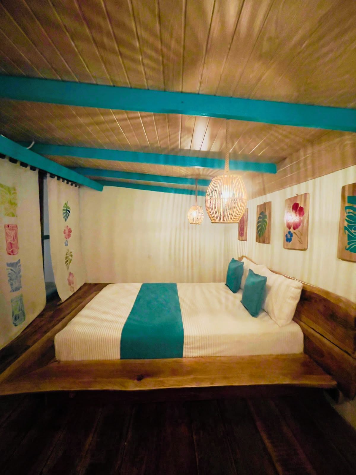 A room with a bed, wooden ceiling beams, a hanging light fixture, and wall art.