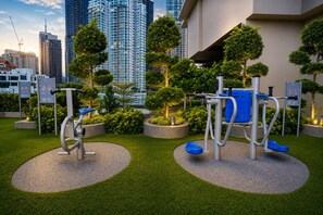 Área de ginástica ao ar livre com equipamentos de exercício, cercada por gramado bem cuidado e árvores, com paisagem urbana ao fundo.