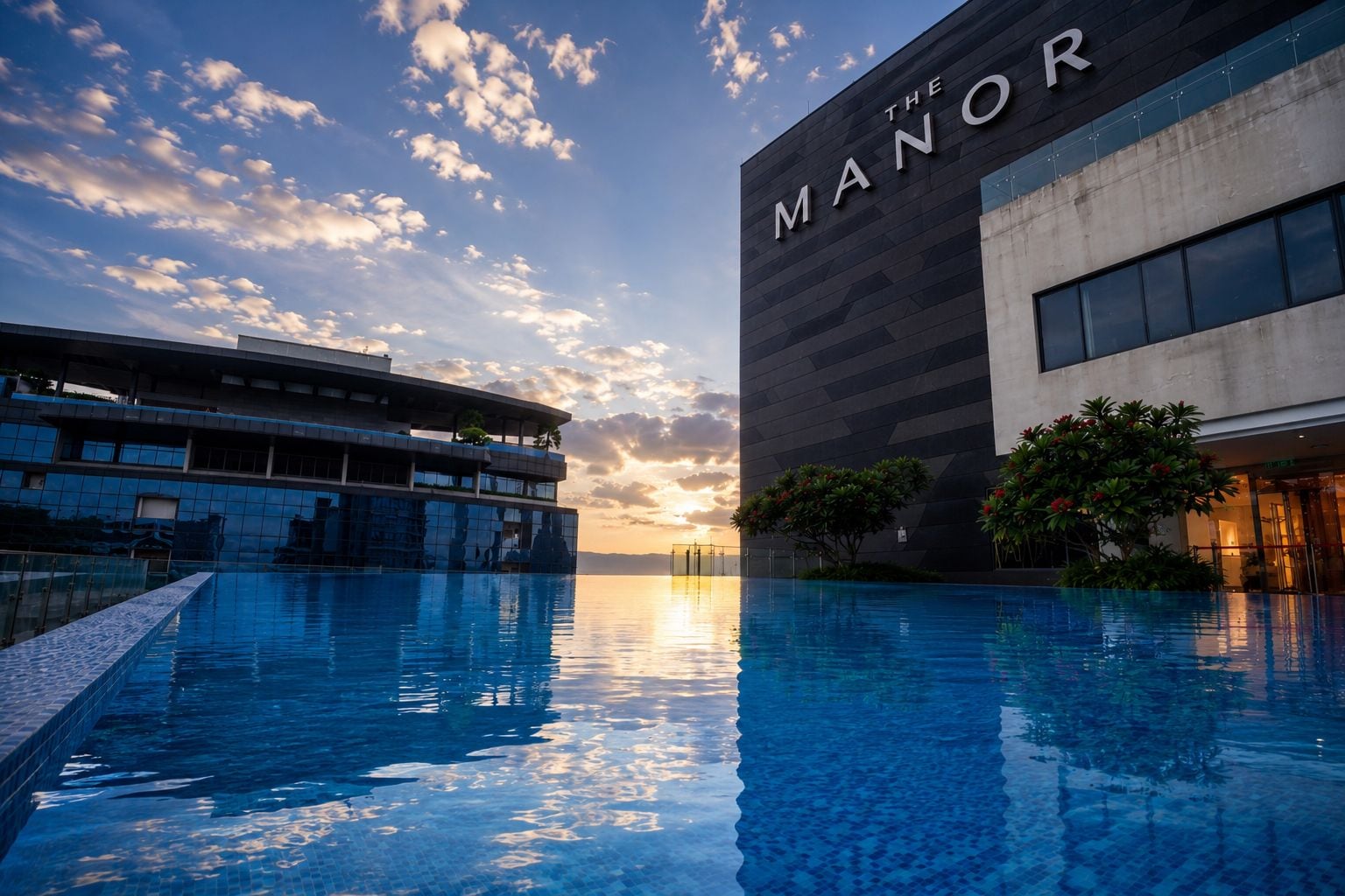 A modern hotel with a large pool, a building with 'THE MANOR' signage, and a sunset sky.