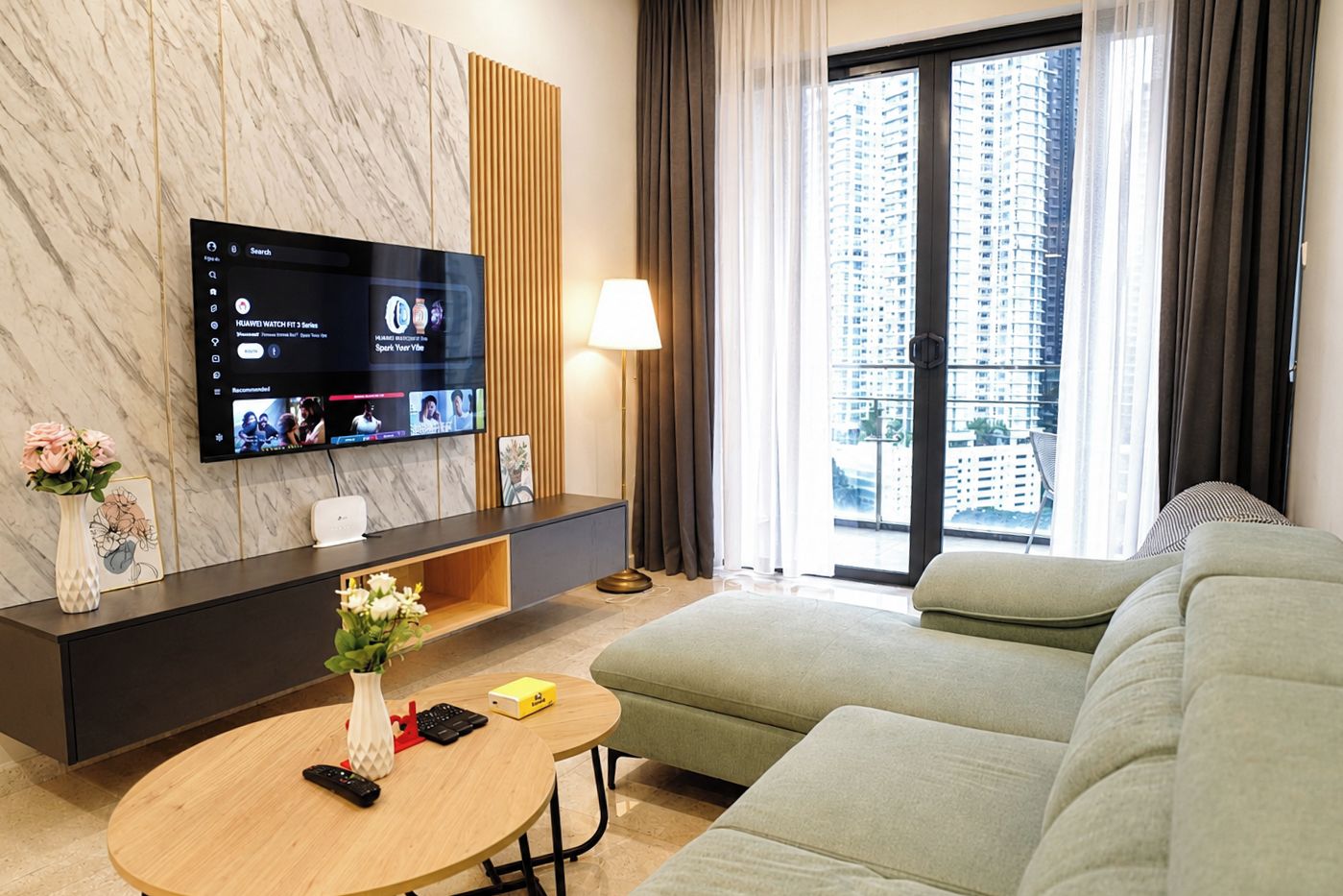 A modern hotel room with a flat-screen TV mounted on a marble wall, a sofa, a coffee table, and a view of a cityscape through large windows.