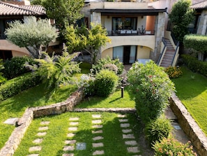 A courtyard with a stone pathway, lush greenery, and a building with a balcony. - Villa Elena Upper Floor – Sea View, Terrace, and Private Garden (San Teodoro)