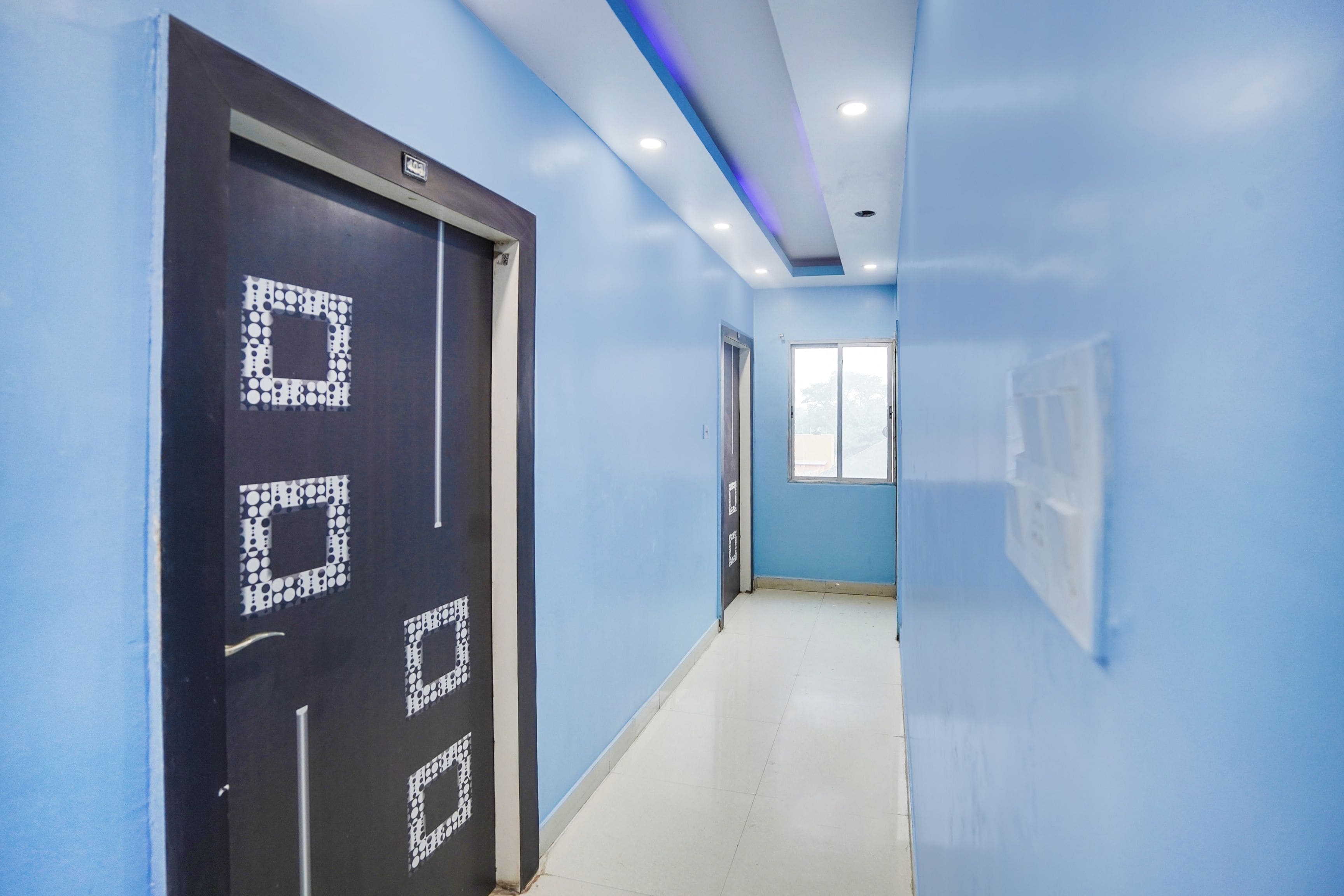 A corridor with blue walls, a black door with a square pattern, and a window at the end.