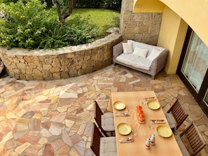 Eine Terrasse im Freien mit Steinboden, einem Esstisch gedeckt mit Tellern, Besteck und Gläsern, sowie einer Korbuschette mit Kissen.