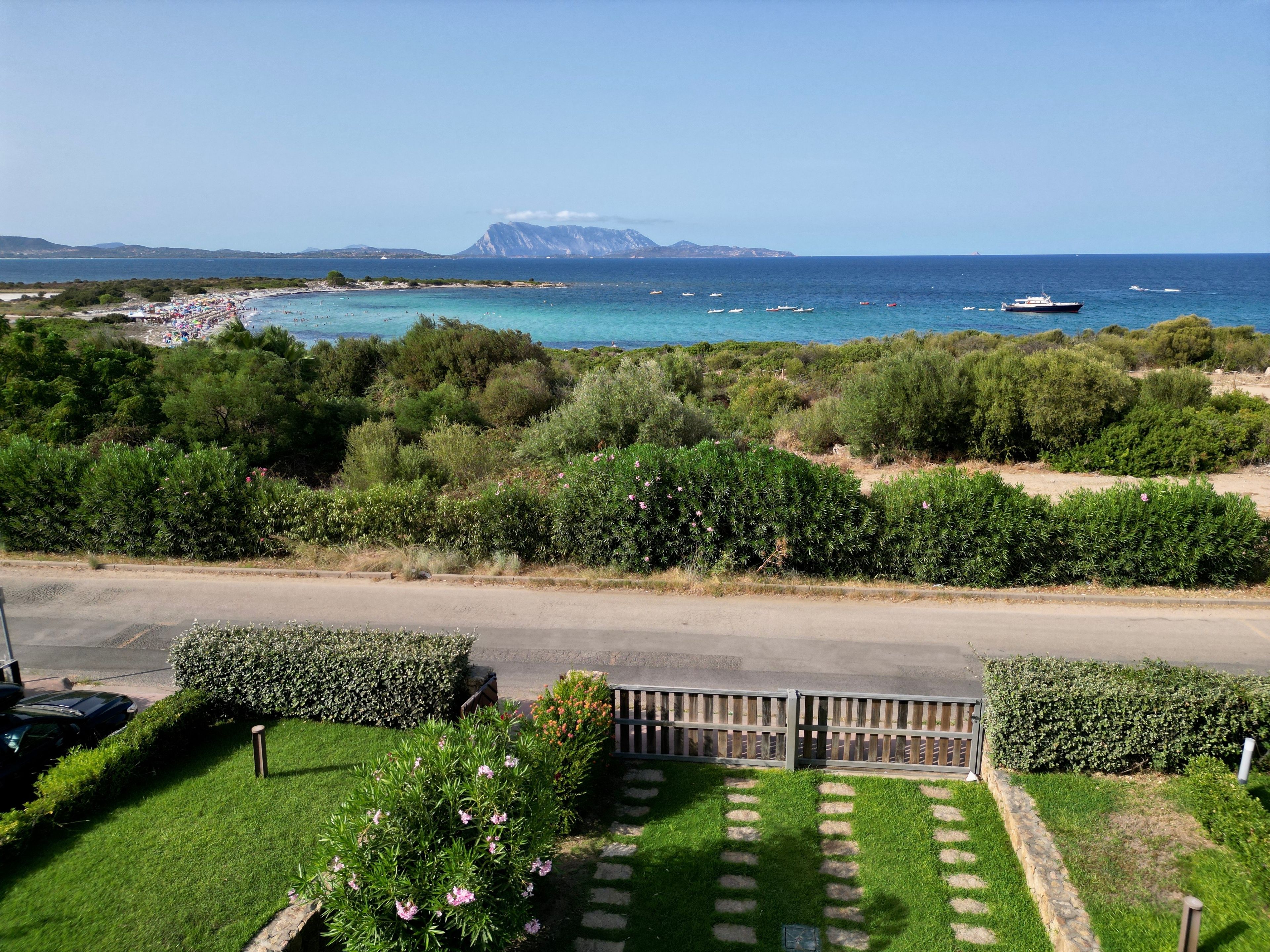 A coastal road with a view of the sea, boats, and a mountain in the distance.