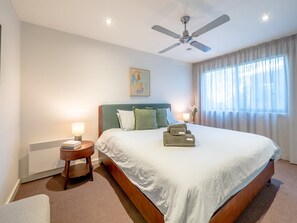 Una habitación de hotel con cama, ventilador de techo, mesita de noche con lámpara y ventana con cortinas.