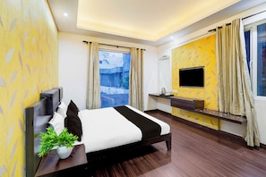 Habitación de hotel con cama, televisor montado en la pared, escritorio pequeño y ventana con cortinas.