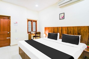 Habitación de hotel con cama, cabecera de madera, escritorio con silla, ventana con rejas y unidad de aire acondicionado.