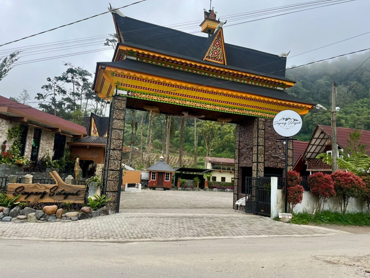 Um portão tradicional com uma placa, entrada pavimentada e montanhas ao fundo.