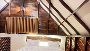 Quarto com teto inclinado, vigas de madeira, cama com roupa de cama branca e janela com cortinas.