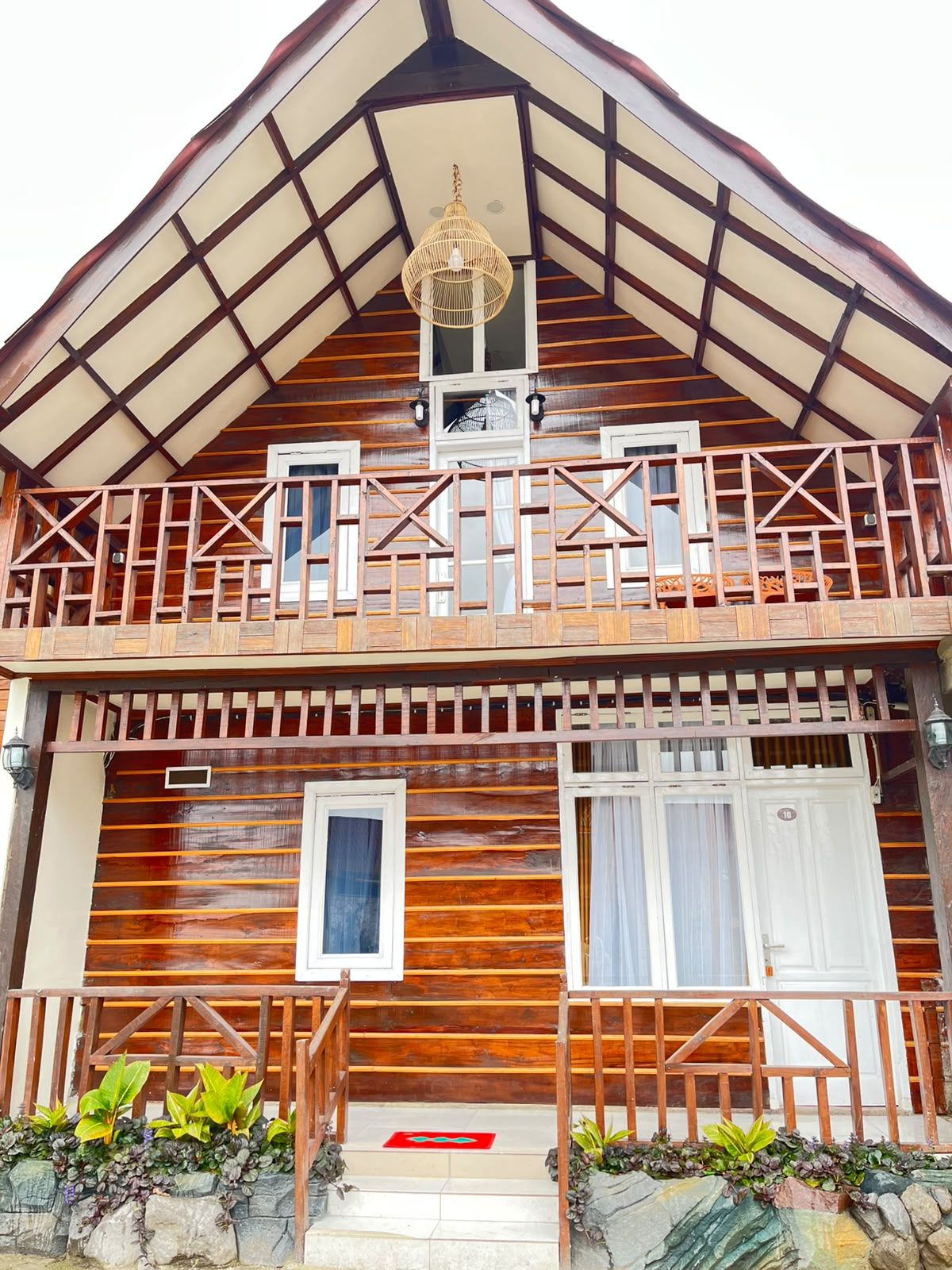 A two-story wooden house with a balcony, a small balcony light, and a red mat at the entrance.
