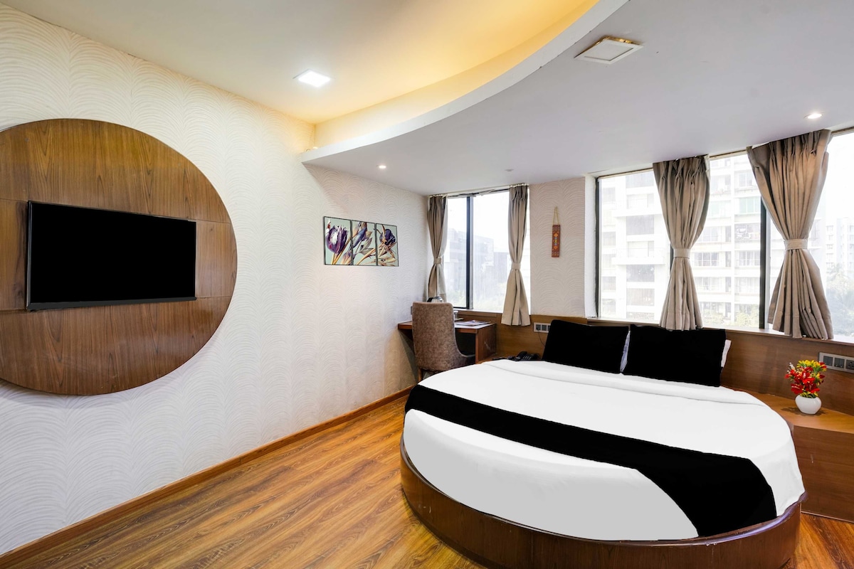 Una habitación de hotel moderna con una cama curvada, un televisor de pantalla plana montado en la pared, un escritorio con una silla y una ventana con cortinas.