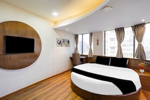 Una habitación de hotel moderna con una cama curvada, un televisor de pantalla plana montado en la pared, un escritorio con una silla y una ventana con cortinas.