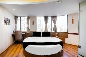 Habitación de hotel con cama redonda, un escritorio con silla y vistas a edificios a través de amplios ventanales.