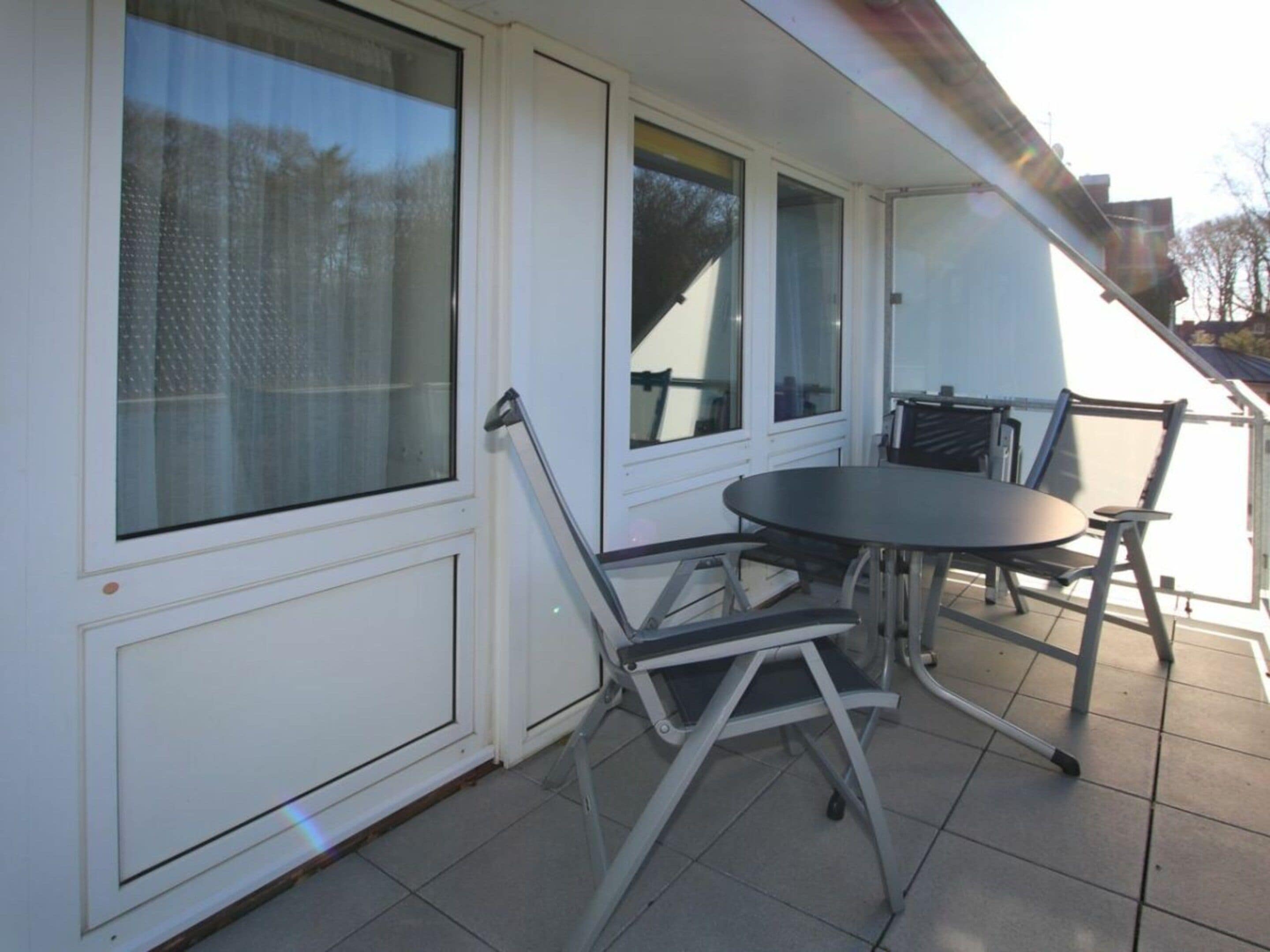 A covered outdoor patio with a table and chairs, large windows, and a white exterior wall.