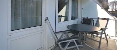 Een overdekt terras buiten met een tafel en stoelen, grote ramen en een witte buitenmuur.