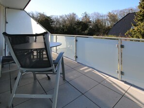 Ein Balkon mit Tisch und Stühlen, eine weiße Wand und Blick auf Bäume sowie ein Gebäude.