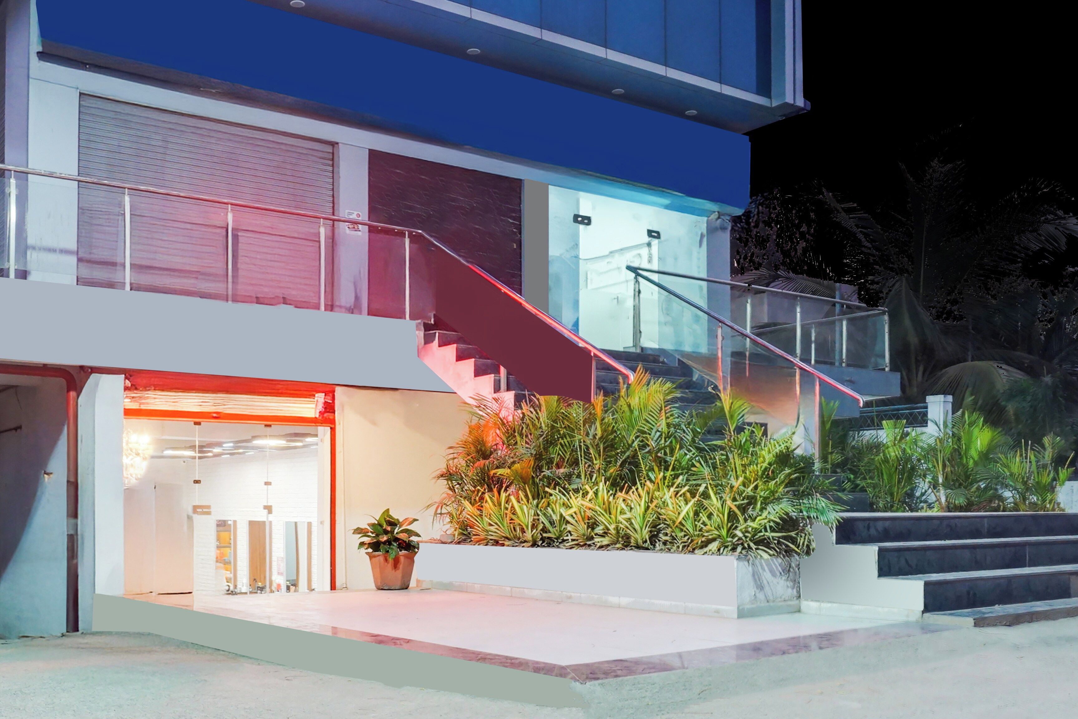 Un bâtiment moderne à deux étages, doté d’une façade en verre, d’un escalier rouge et d’un petit jardin agrémenté de plantes en pot.